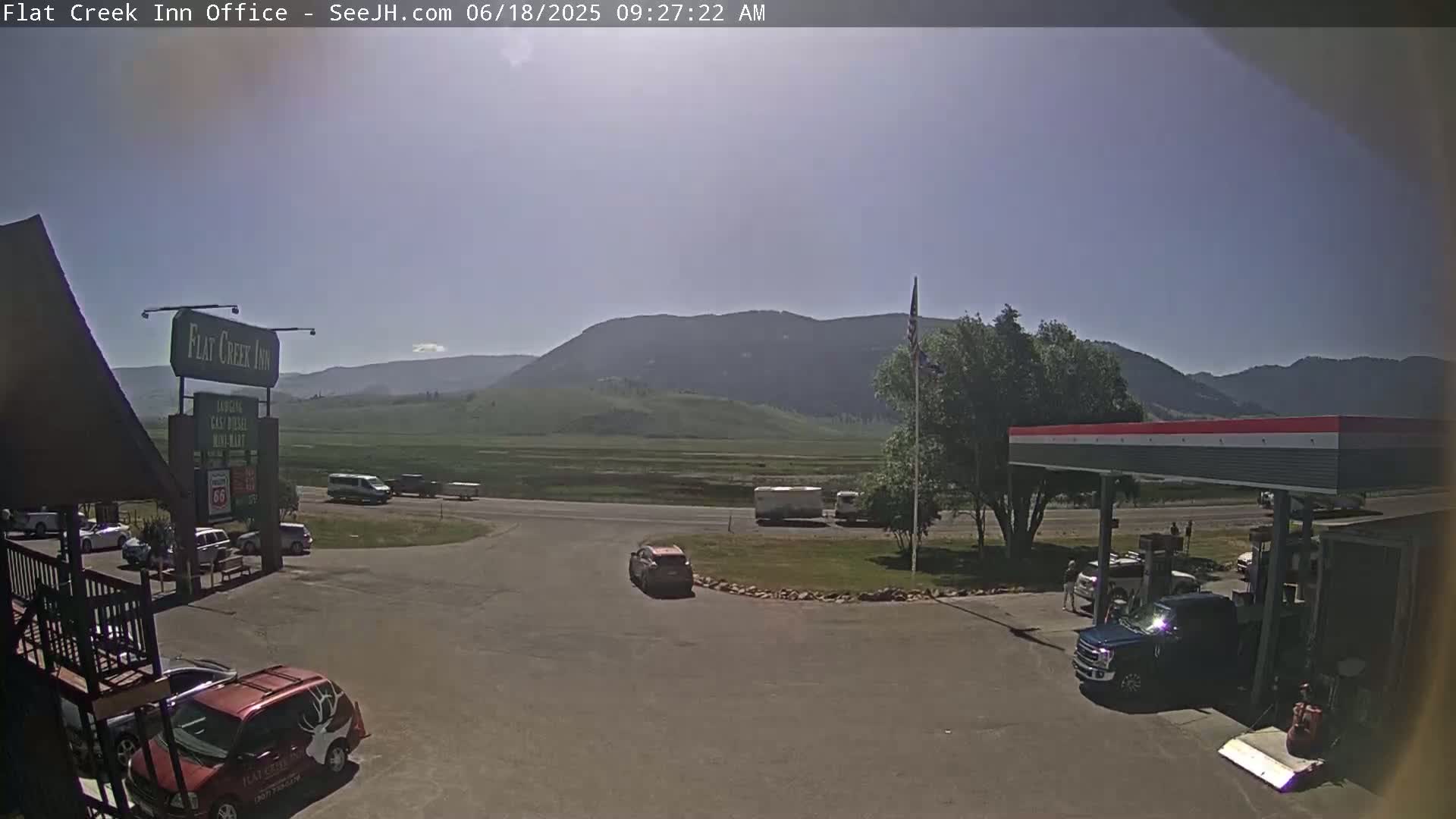 A sunny, mostly clear day reveals a gas station and inn parking lot with several vehicles, situated before a vast, flat plain and distant mountains.