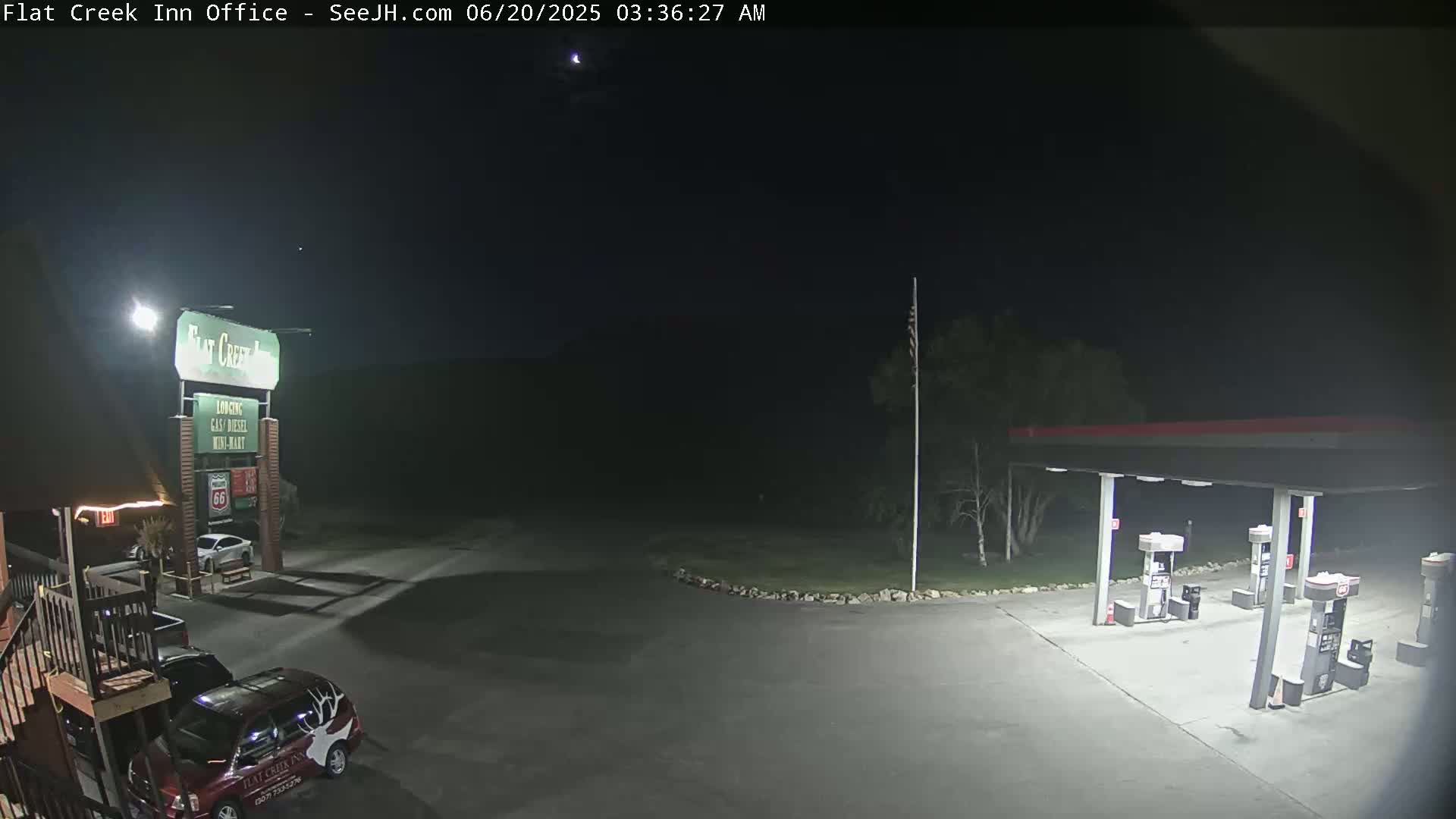 A nighttime view of a gas station and motel parking lot under a clear, dark sky, illuminated by artificial lights.