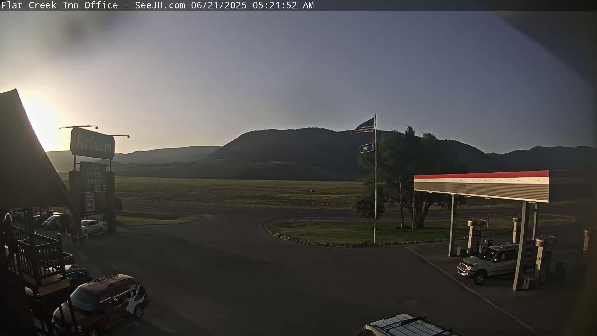 A gas station and inn are situated on a flat expanse of land before a range of dark mountains, under a clear, dusky sky at dawn.