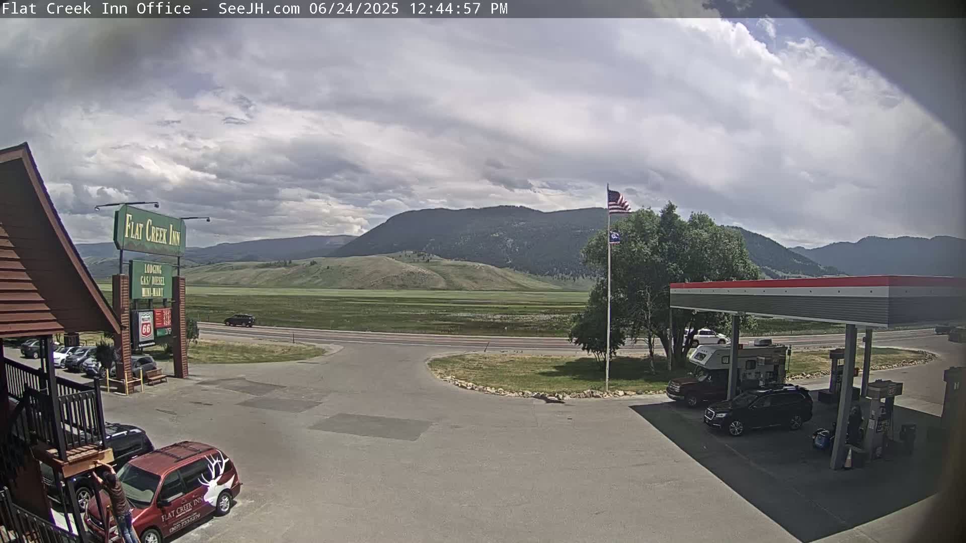 A partly cloudy day shows a gas station and motel parking lot with vehicles, set against a backdrop of rolling green hills and mountains.