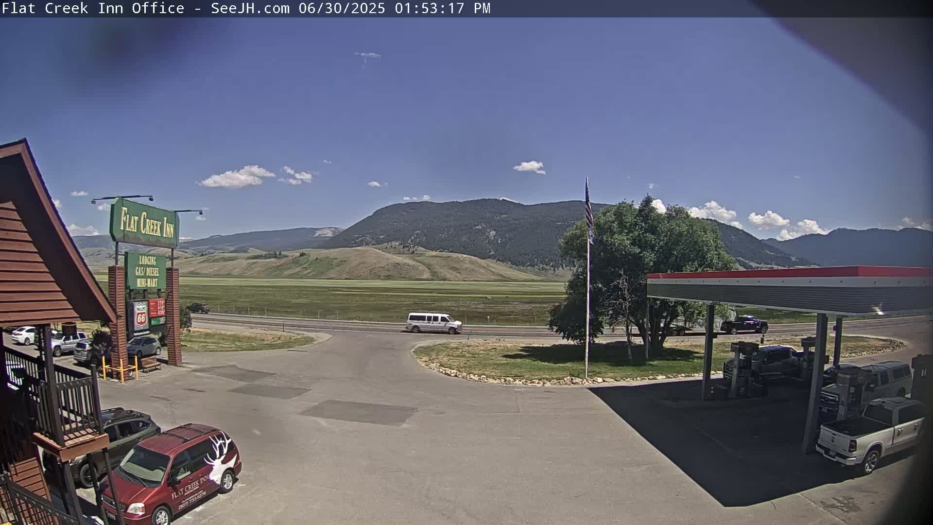 A gas station and inn are situated in a parking lot, with a van driving on a road in front of mountains under a mostly sunny sky.