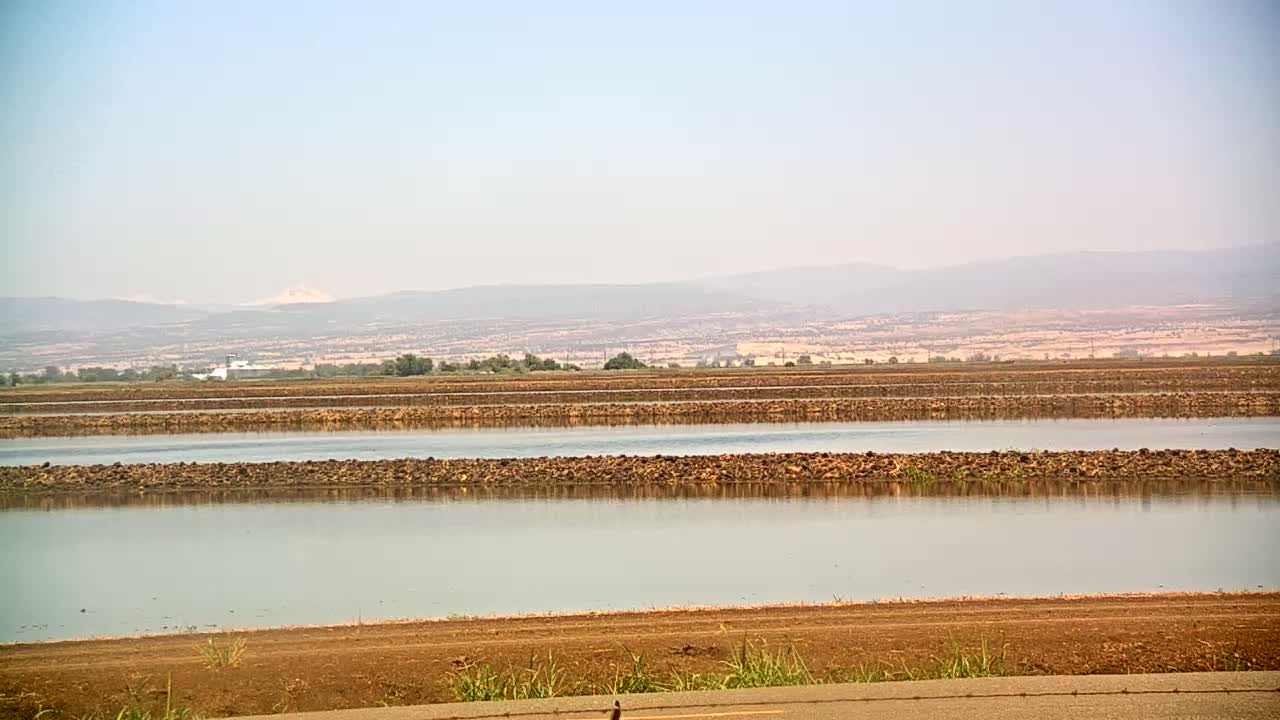 Under a hazy sky, calm water fills several rectangular basins separated by low earthen walls in a vast flat agricultural landscape, with distant mountains visible on the horizon.