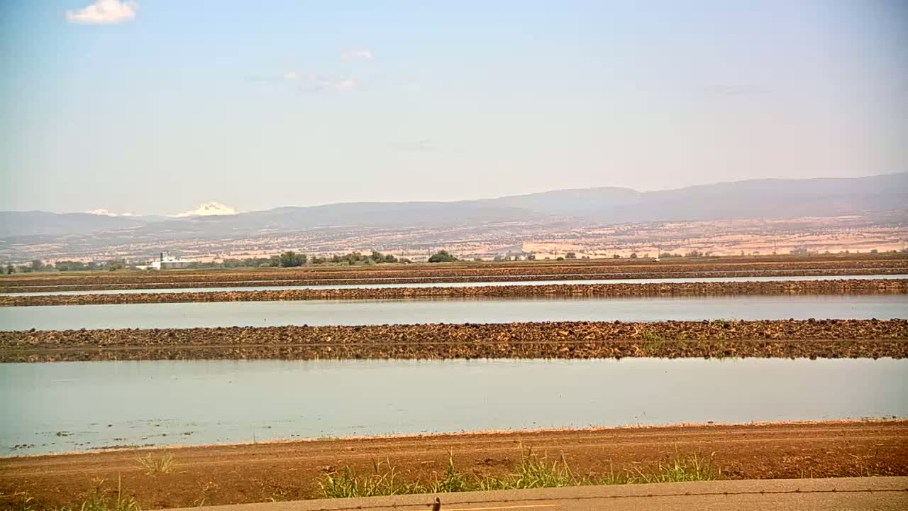 Calm, flat water in several long rectangular sections separated by low, dark brown barriers, stretches across a flat landscape under a mostly clear sky, with mountains visible in the distance.