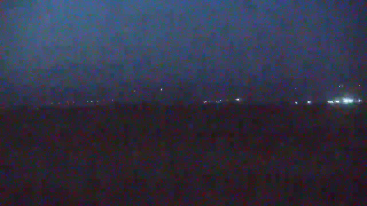 A dark, hazy night scene shows a distant line of faint lights on the horizon above a dark, flat landscape.