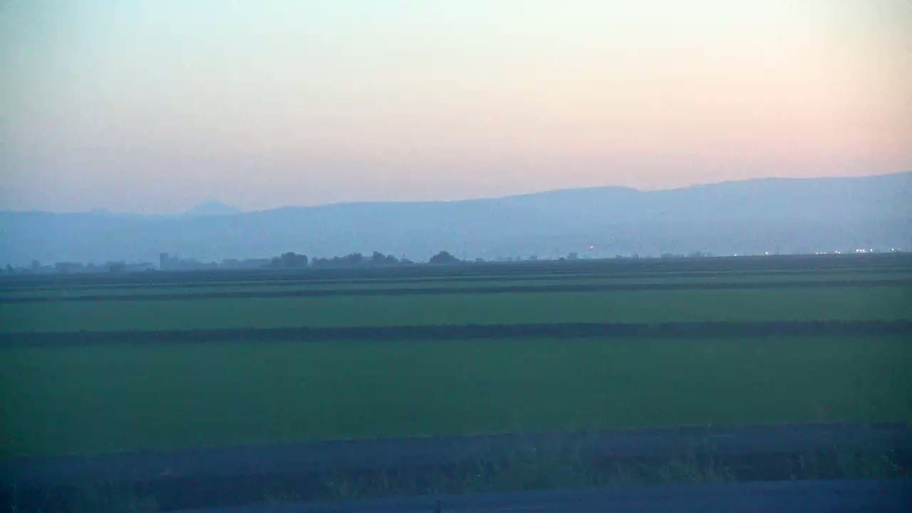 A hazy, pale-pink sunset illuminates a flat, green agricultural landscape with distant, bluish mountains.