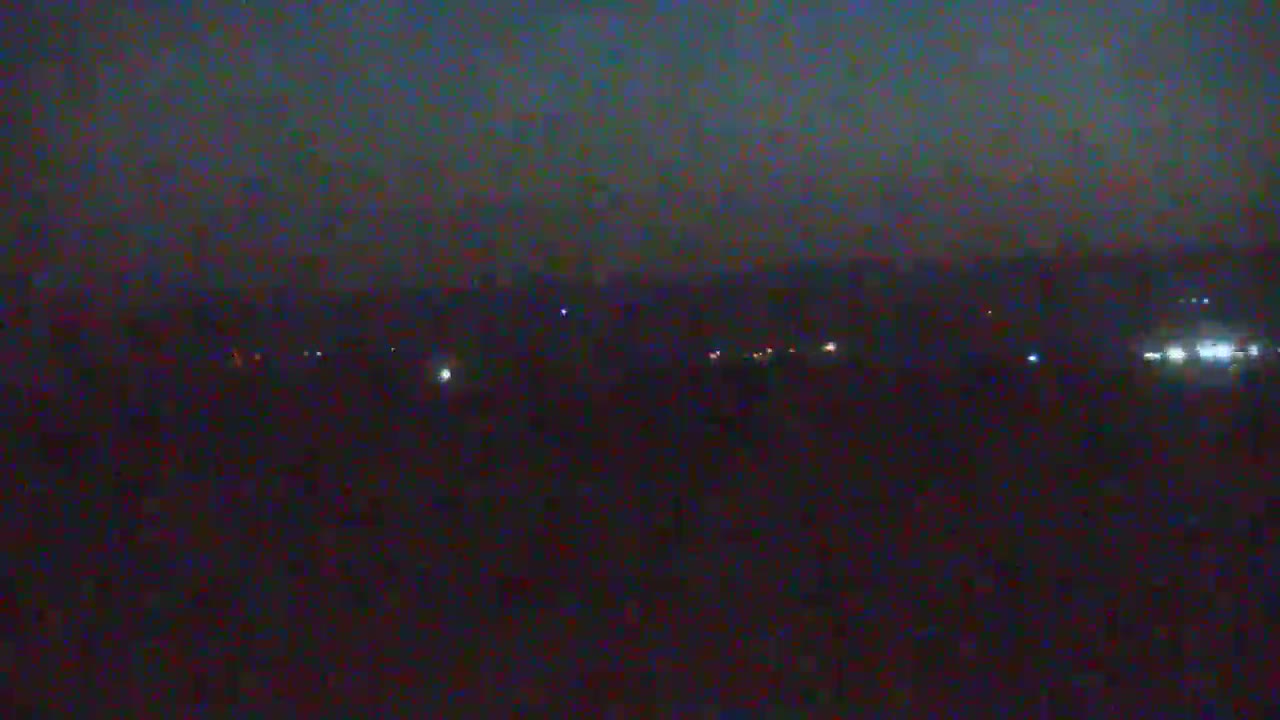 A nighttime long shot shows a dark landscape with a few faint lights in the distance under a dark sky.