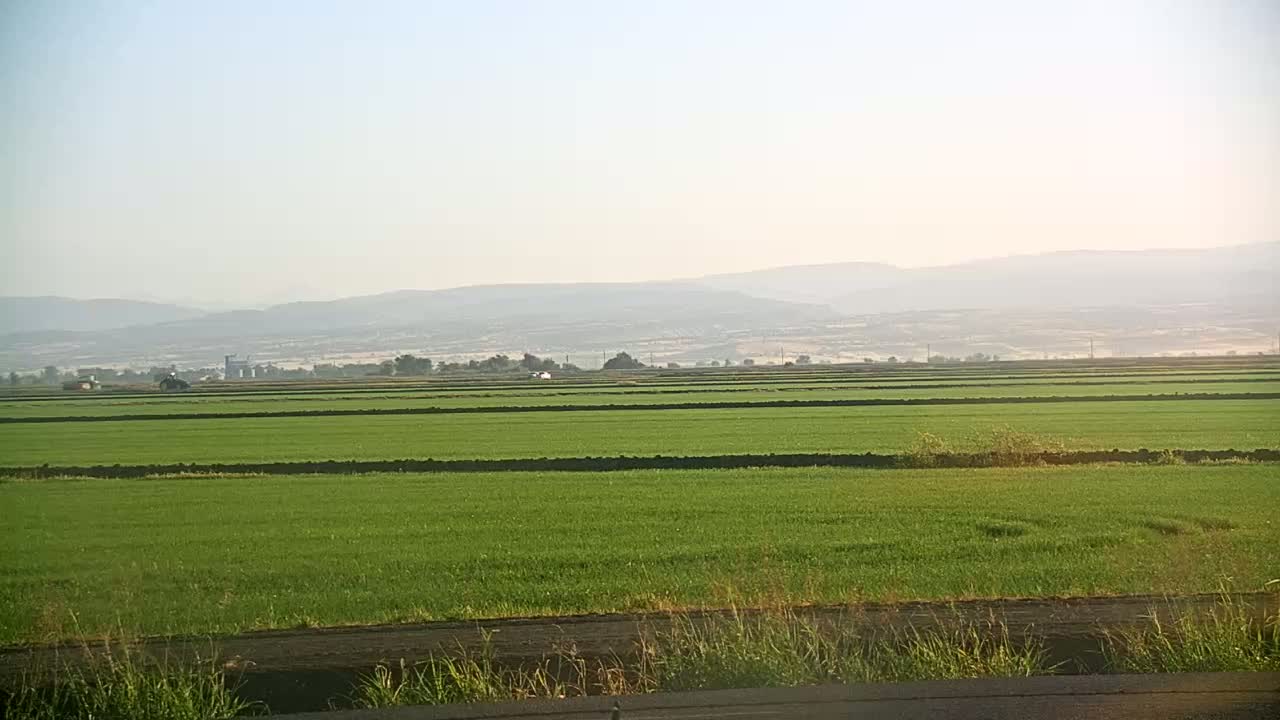 A vast expanse of bright green fields stretches to a hazy mountain range under a clear, light sky.