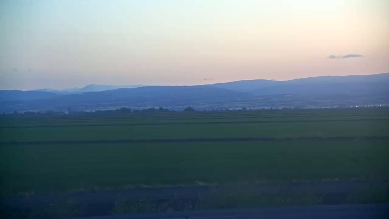 A vast, flat expanse of green fields stretches toward a hazy, blue-toned mountain range under a pale sunset sky.