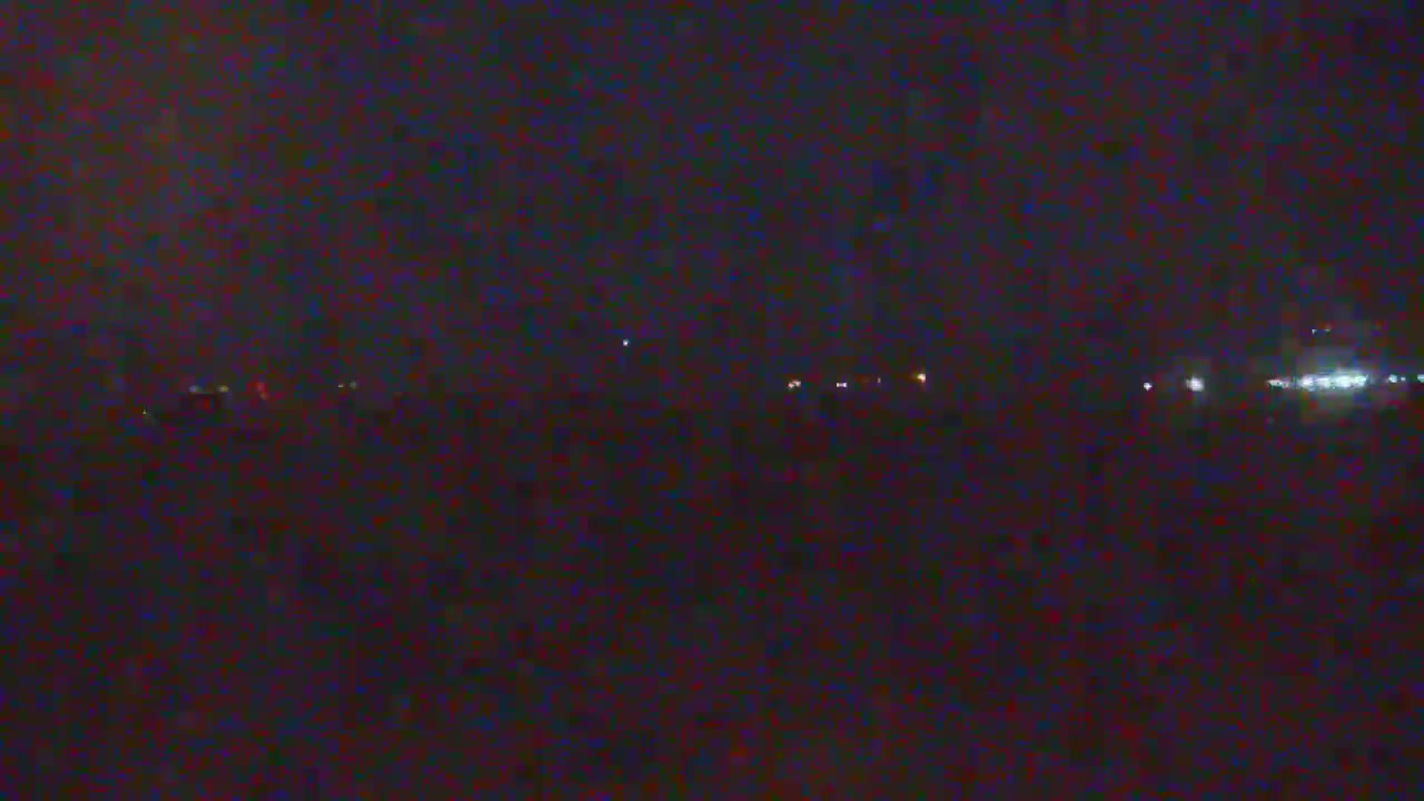 A dark, nighttime scene shows a distant line of faint lights across a flat, dark landscape under a mostly clear sky.