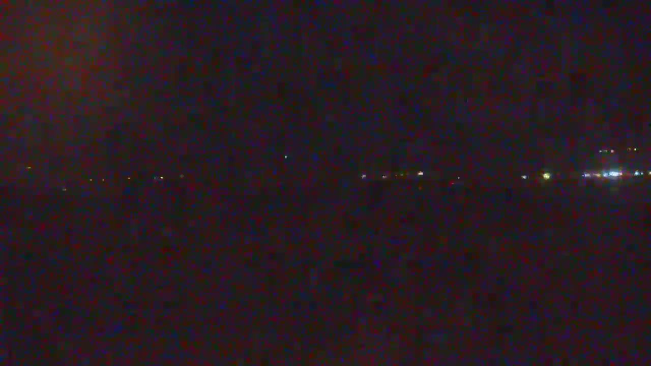 A dark, nighttime scene shows a distant line of faint lights across a flat, dark landscape under a mostly clear sky.
