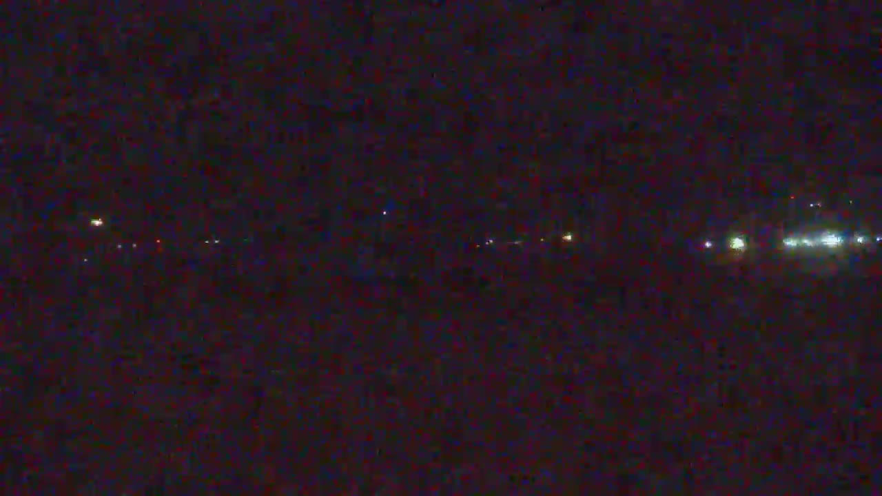 A dark, nighttime scene shows a distant line of faint lights across a flat, dark landscape under a mostly clear sky.