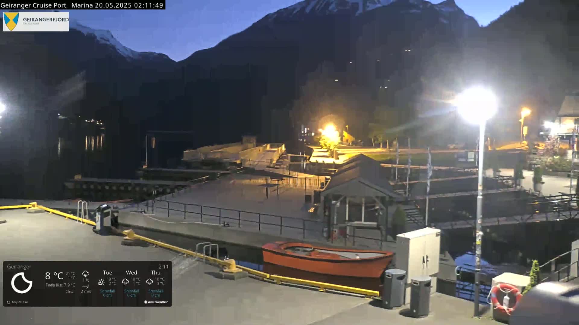 A nighttime view of a marina nestled at the base of dark mountains under a clear sky shows a small red boat and various dockside structures.