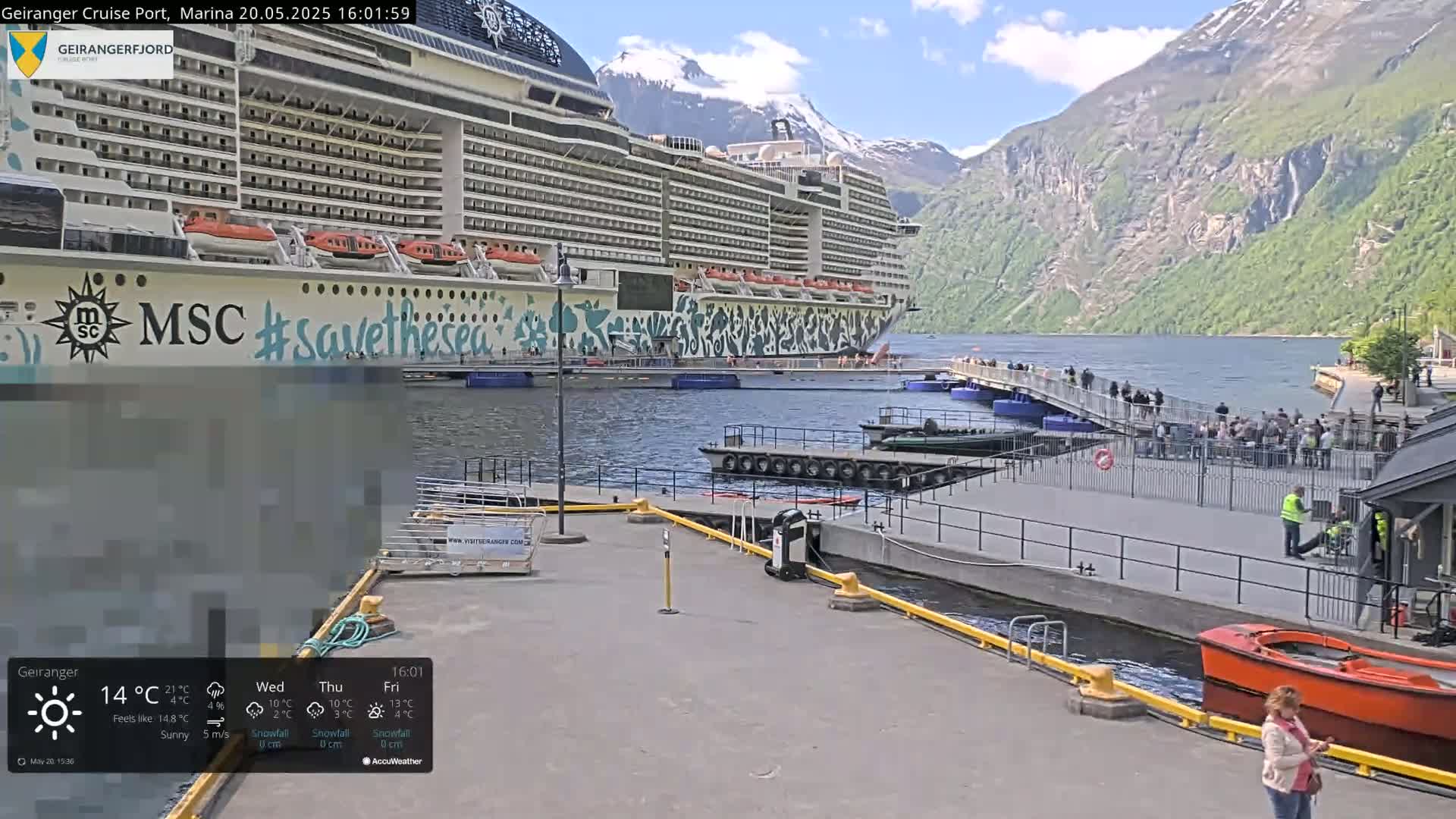 A large cruise ship is docked at a port, with people walking on a pier next to it; mountains and calm water are in the background on a sunny day.