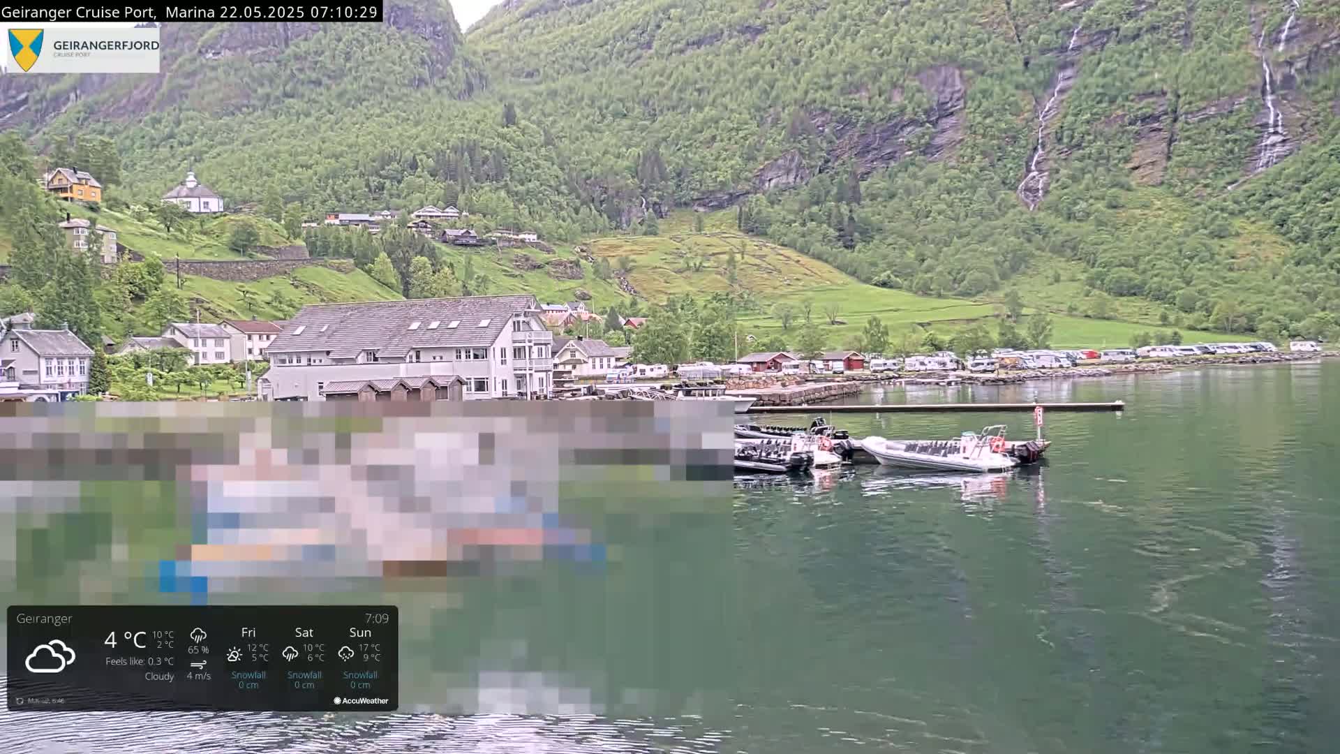 A cloudy day in a fjord town shows a calm body of water with several boats docked near a waterfront building, backed by a hillside village and steep green mountains with waterfalls.