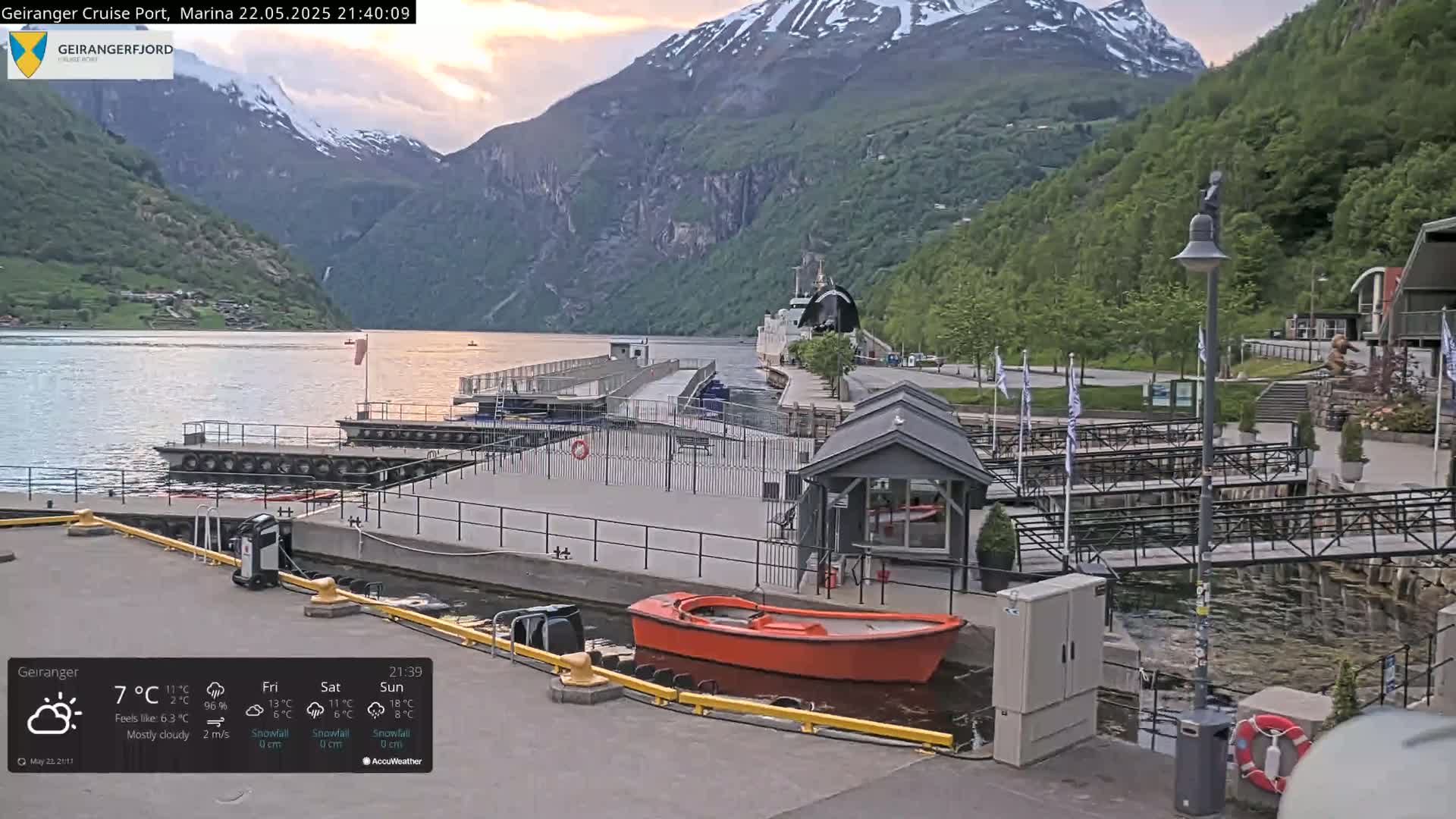 A calm harbor scene at dusk, nestled between steep, snow-capped mountains, with a small orange boat moored near a dock.