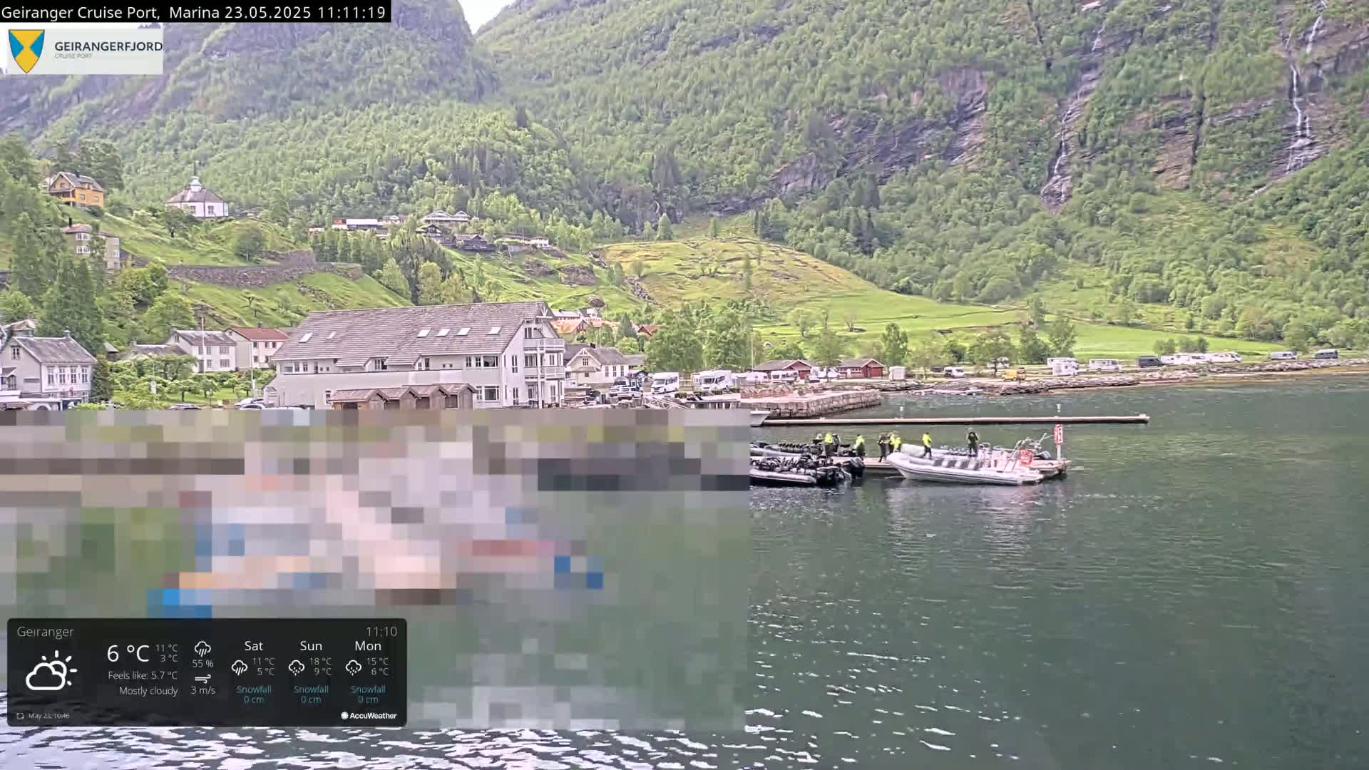 A mostly cloudy day shows a fjordside village with several small boats at a dock nestled between steep, green mountains.