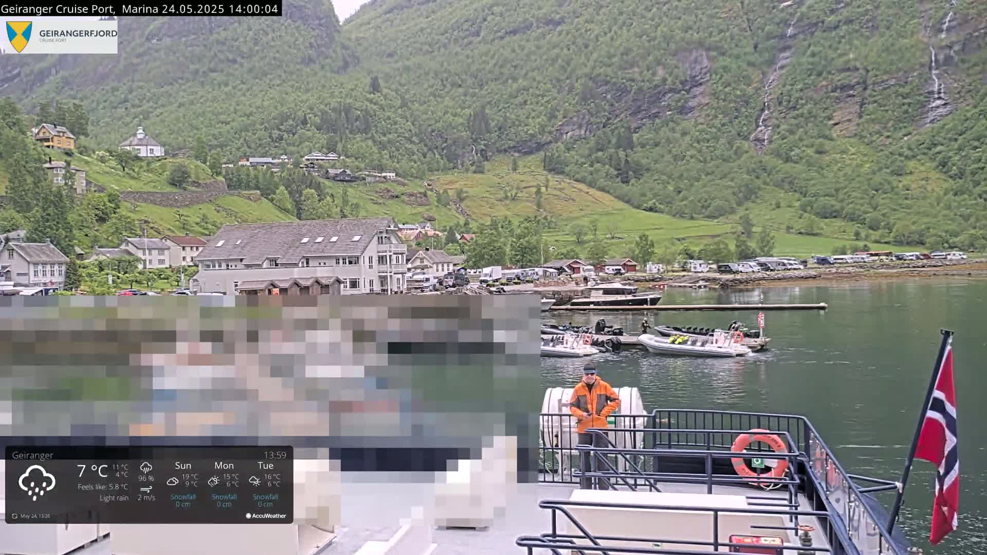 A rain-slicked harbor scene shows a village nestled against steep, green mountains, with several boats moored at a dock and a person standing on the deck of a larger vessel in the foreground.