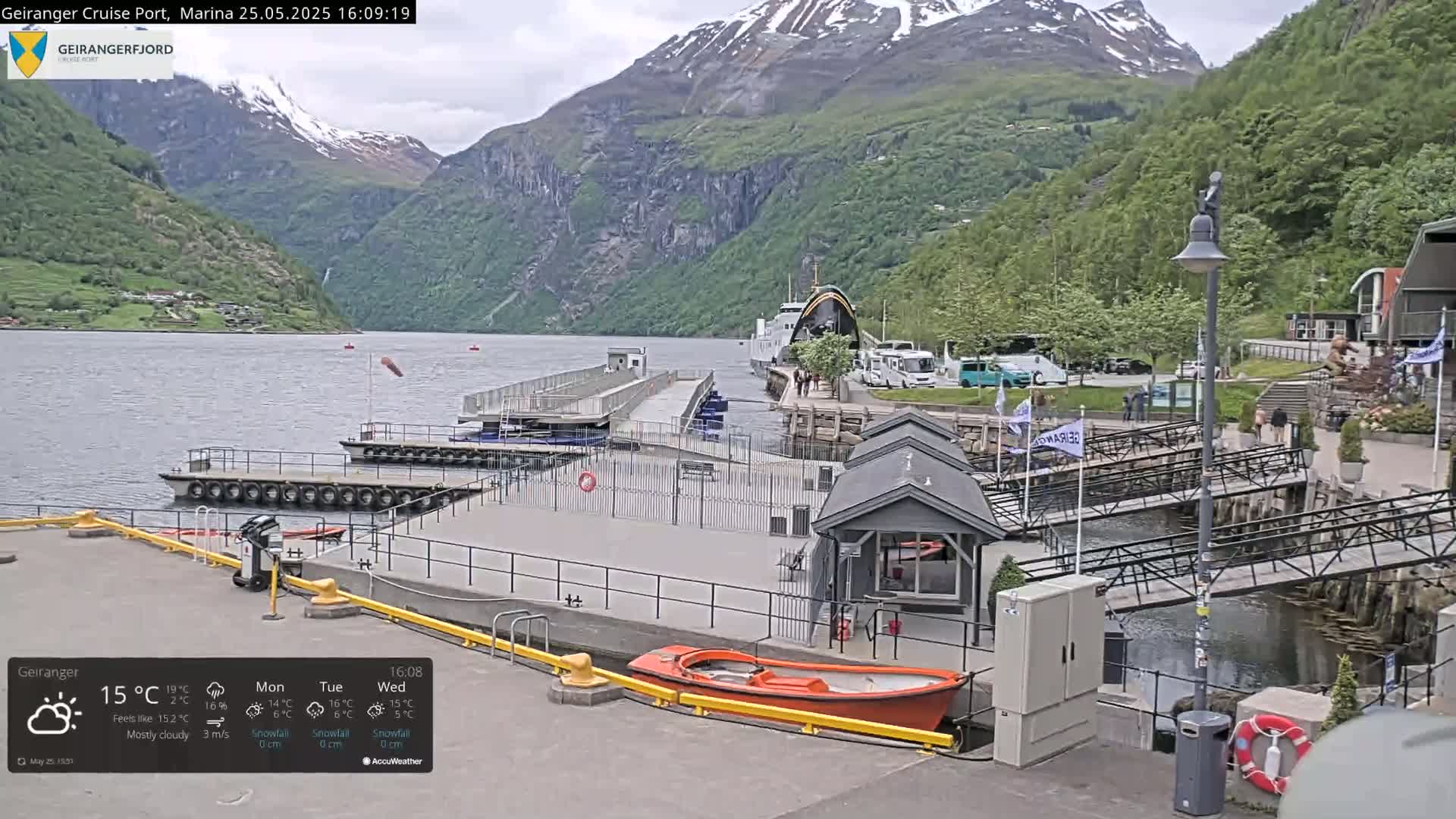 A mostly cloudy day at a marina nestled between steep, snow-capped mountains, with a ferry docked and small boats moored nearby.
