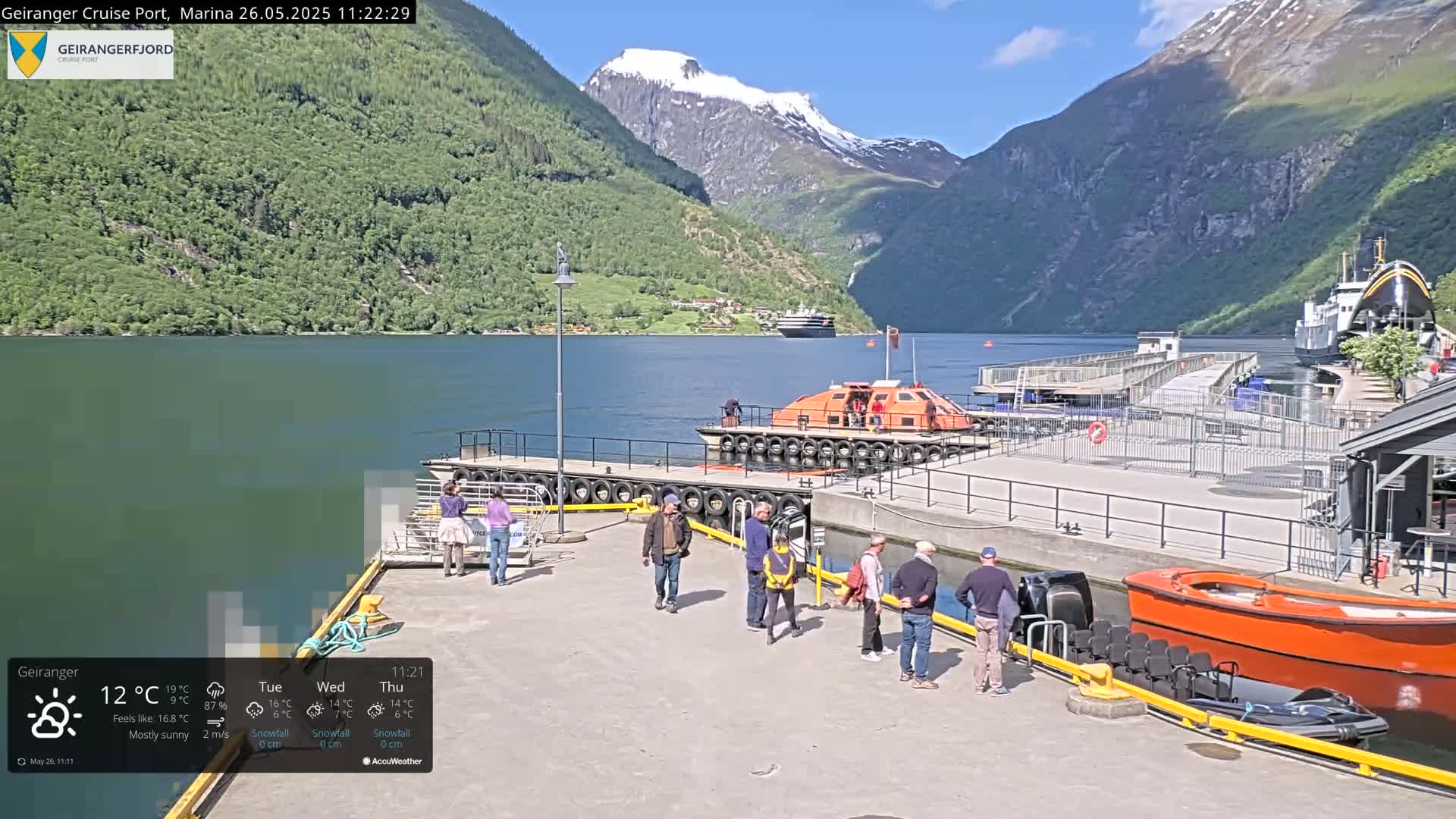 A sunny day in a fjord, showing a cruise ship in the distance, several small boats at a dock, and people gathered on the dock.