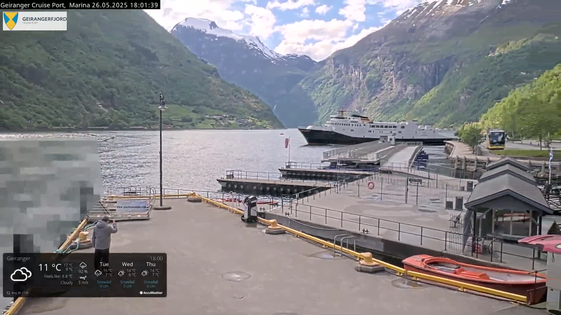 A cruise ship is docked at a marina in a fjord surrounded by steep, verdant mountains under a partly cloudy sky.