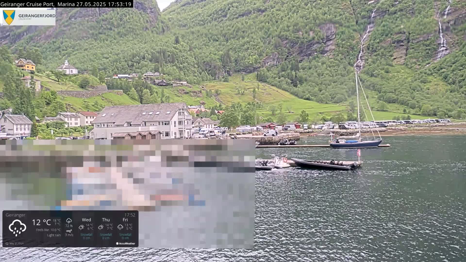 A calm body of water, with several boats docked near a small village nestled at the base of steep, green mountains, under light rain.