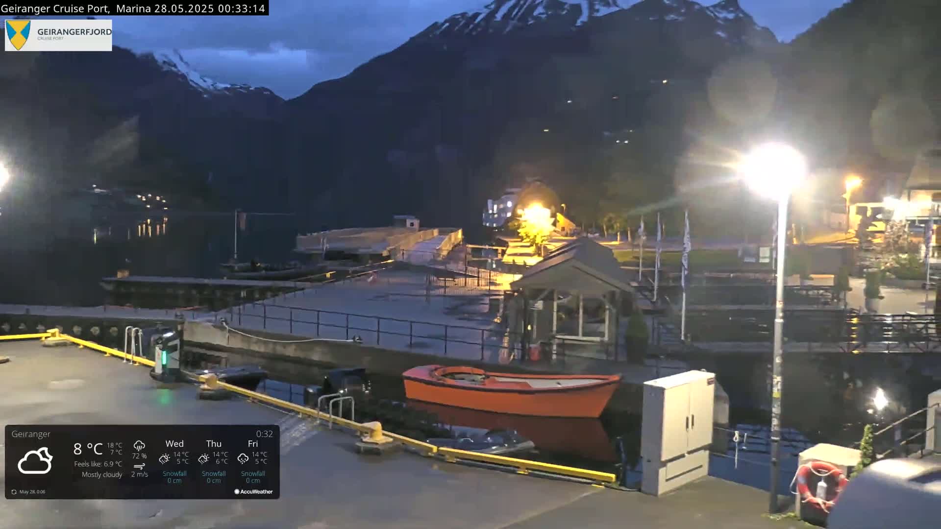 A mostly cloudy night scene shows a marina with a small orange boat in the water, near a dock and buildings, with mountains in the background.