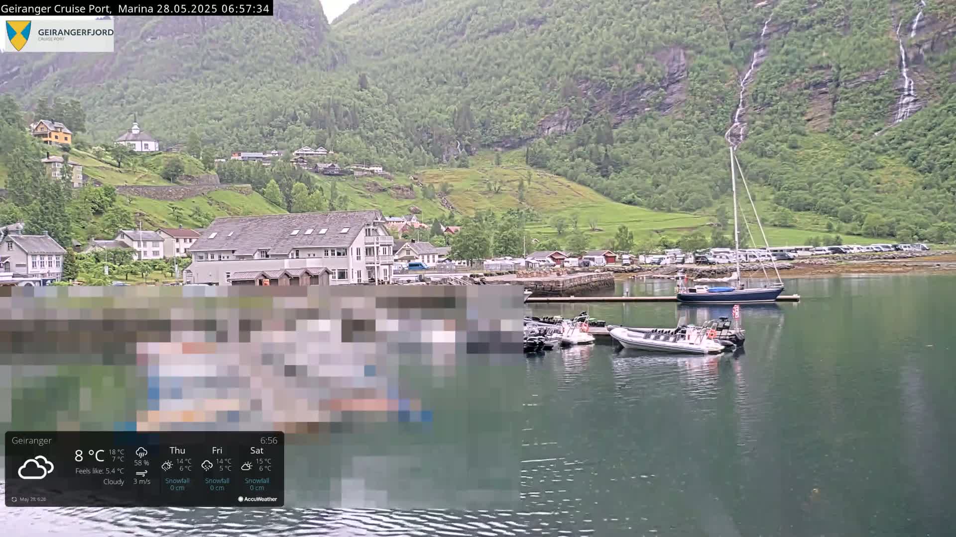 A cloudy day in a harbor town nestled at the base of a verdant, mountainous fjord, with several boats moored and a sailboat at anchor.