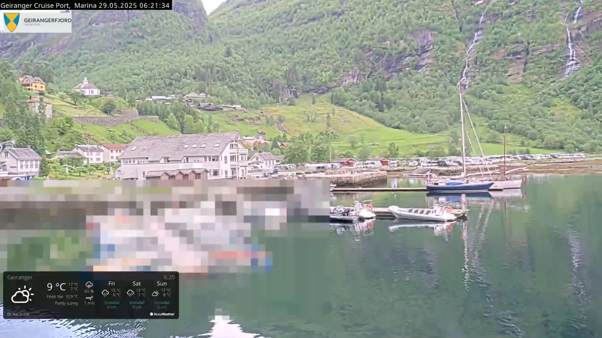 A partly sunny waterfront village nestled against a verdant mountainside with waterfalls cascading down, features several boats moored in a calm harbor.