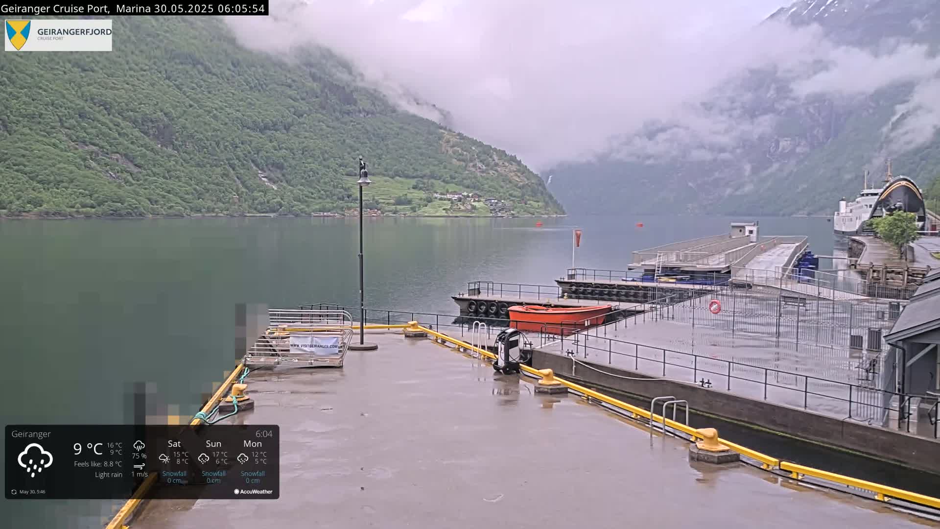 A cruise ship is docked at a marina on a fjord surrounded by mountains under a light rain.