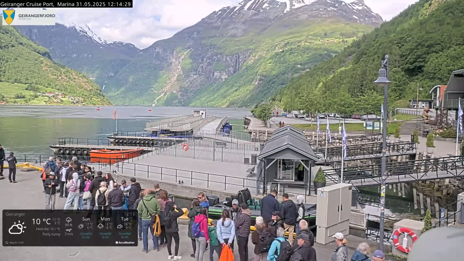 A crowd of people stand near a dock in a fjord surrounded by steep, green mountains under partly sunny skies.