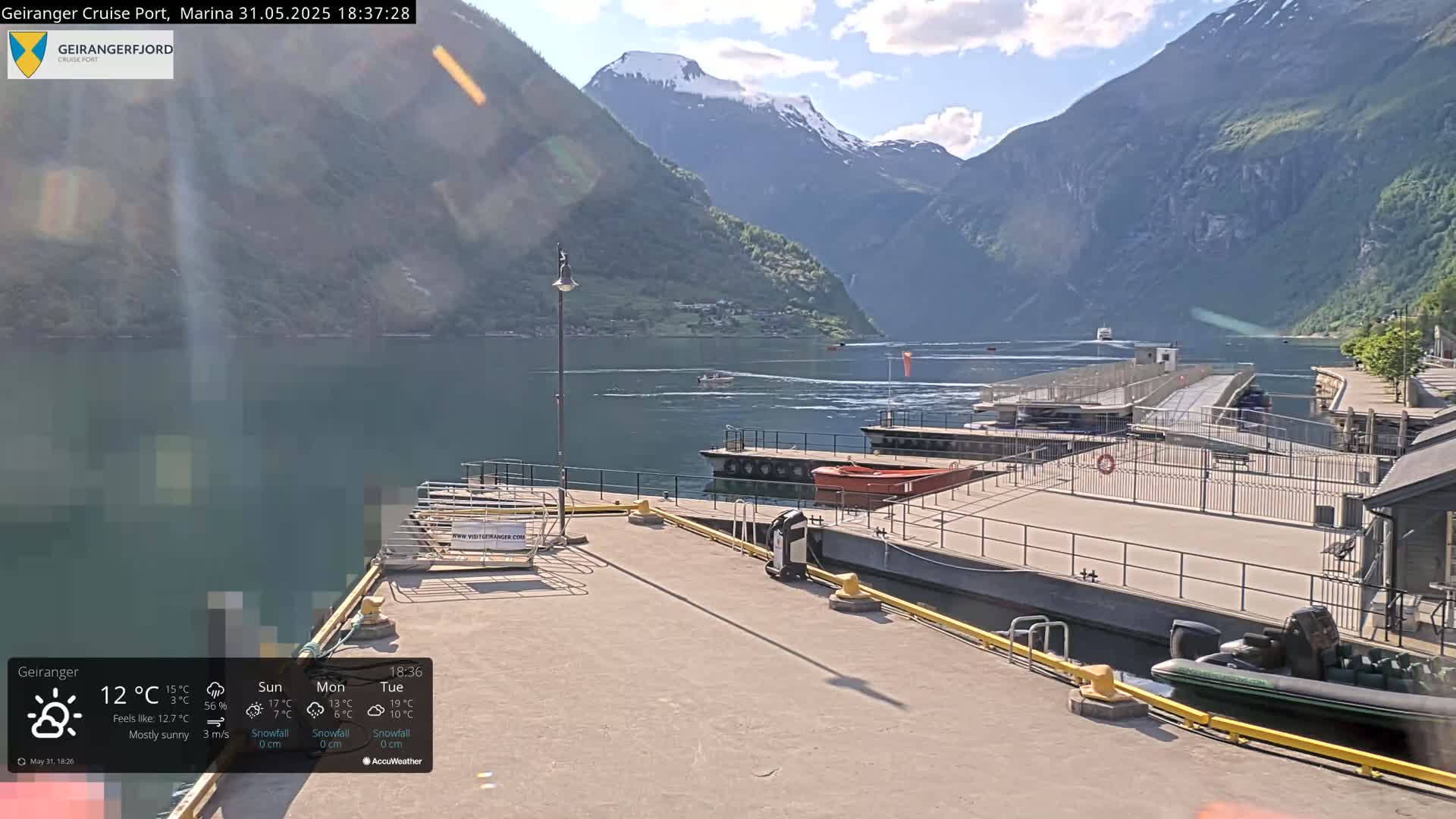 A mostly sunny view of a marina with several boats docked, situated in a fjord surrounded by steep, green mountains, one of which has a snow-capped peak.