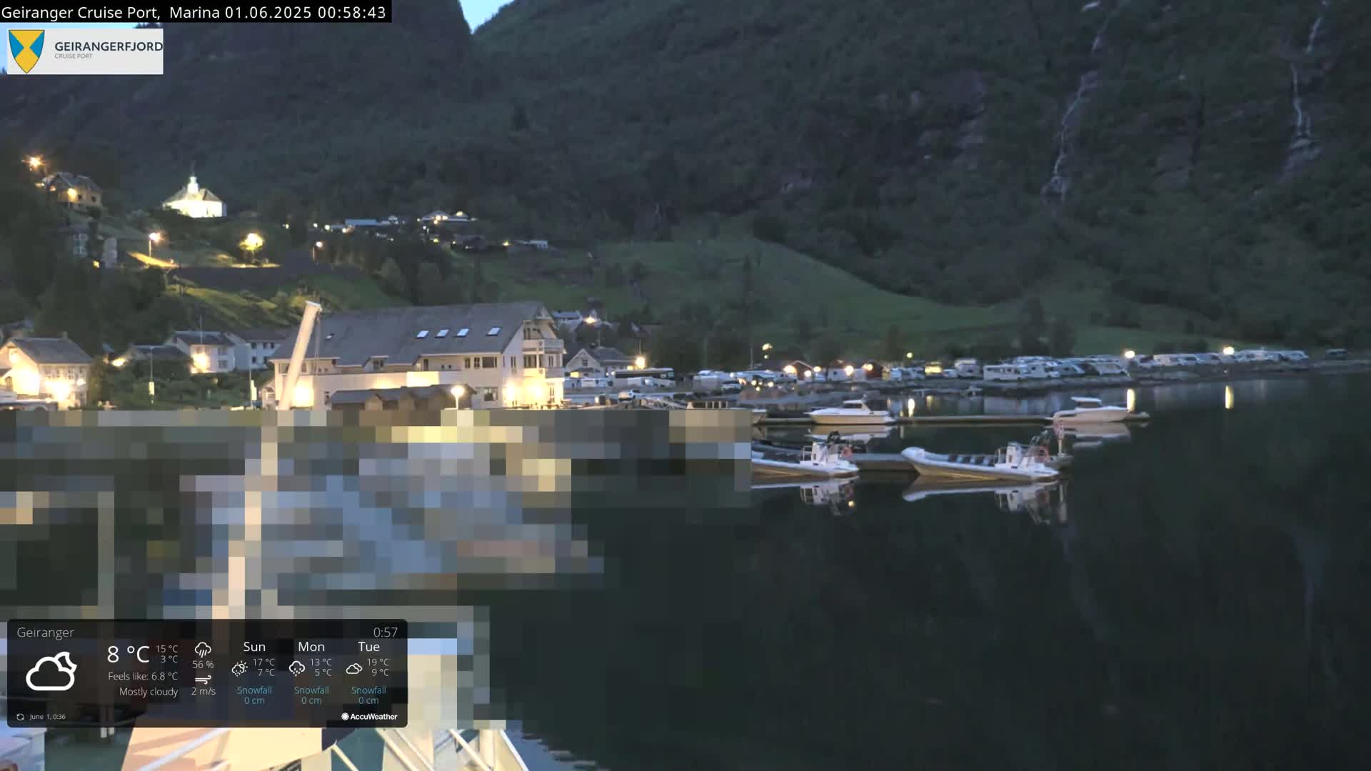 A mostly cloudy night scene shows a small waterfront town nestled beside a fjord, with several boats moored in calm water at the base of dark, steep hills.