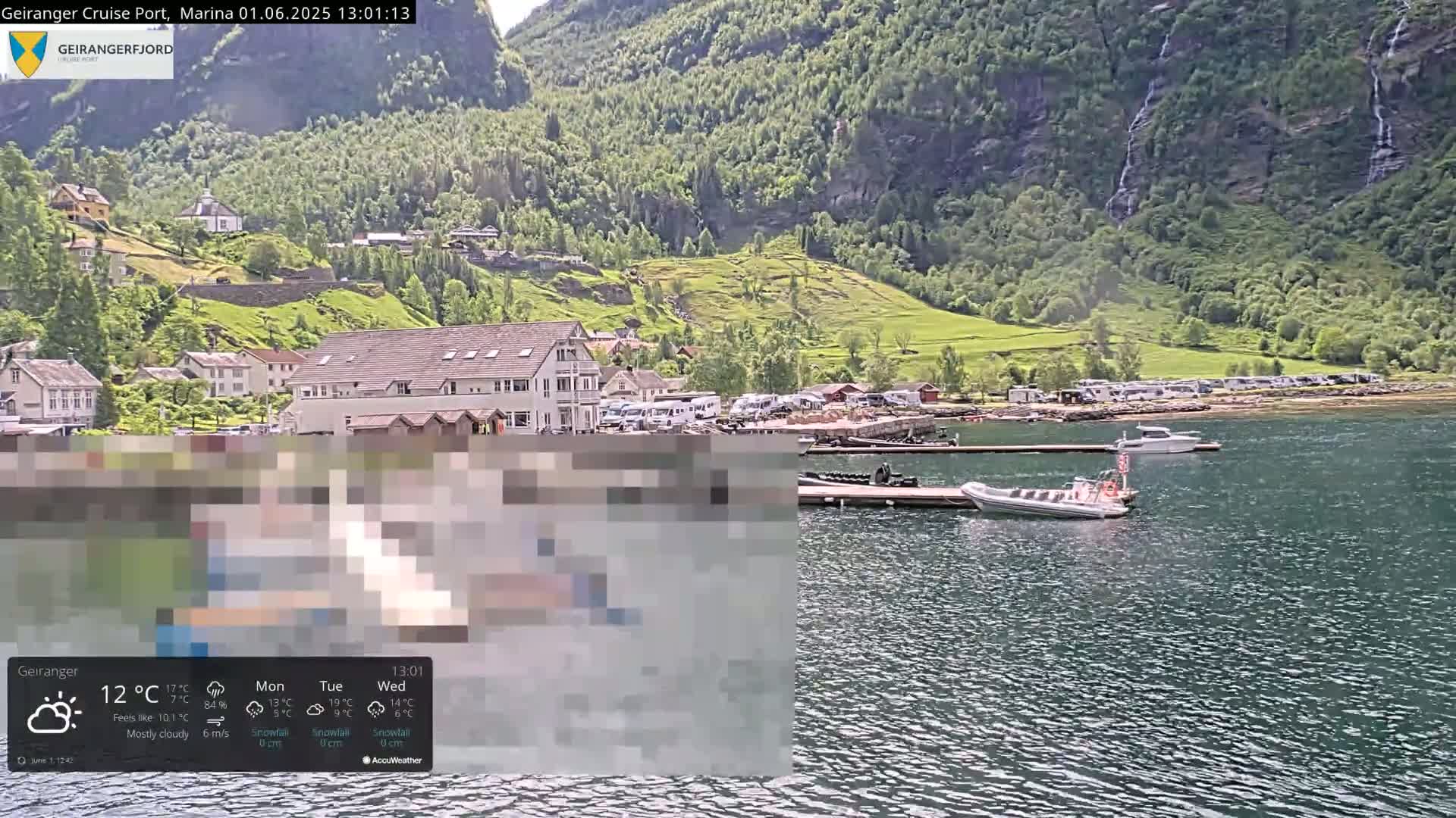 A mostly cloudy day at a marina nestled in a fjord, with several boats docked and RVs parked on the shore, backed by green hills and waterfalls cascading down a mountainside.