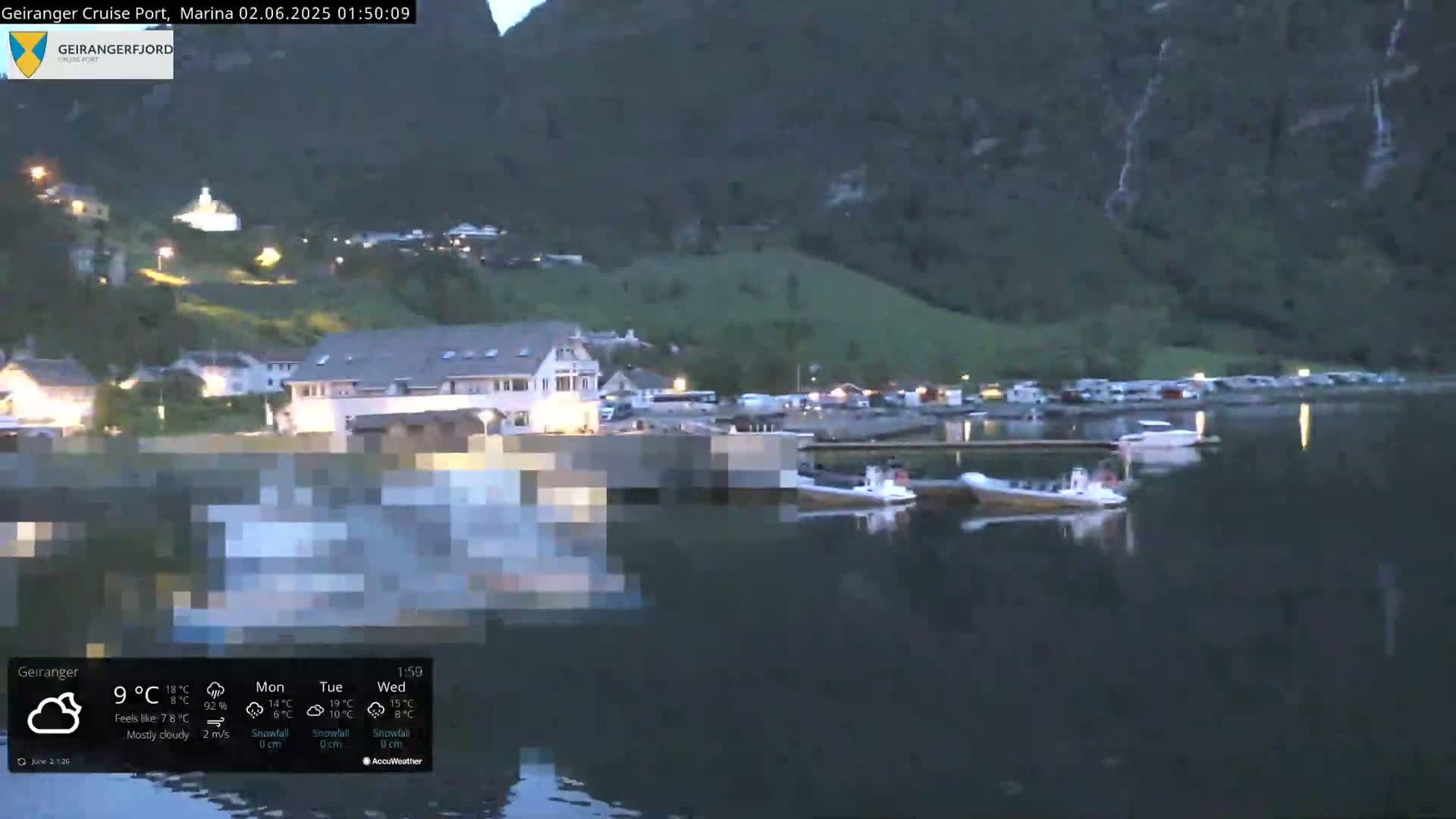 A mostly cloudy night scene shows a small village nestled at the base of steep, dark mountains beside a calm body of water with several small boats moored.