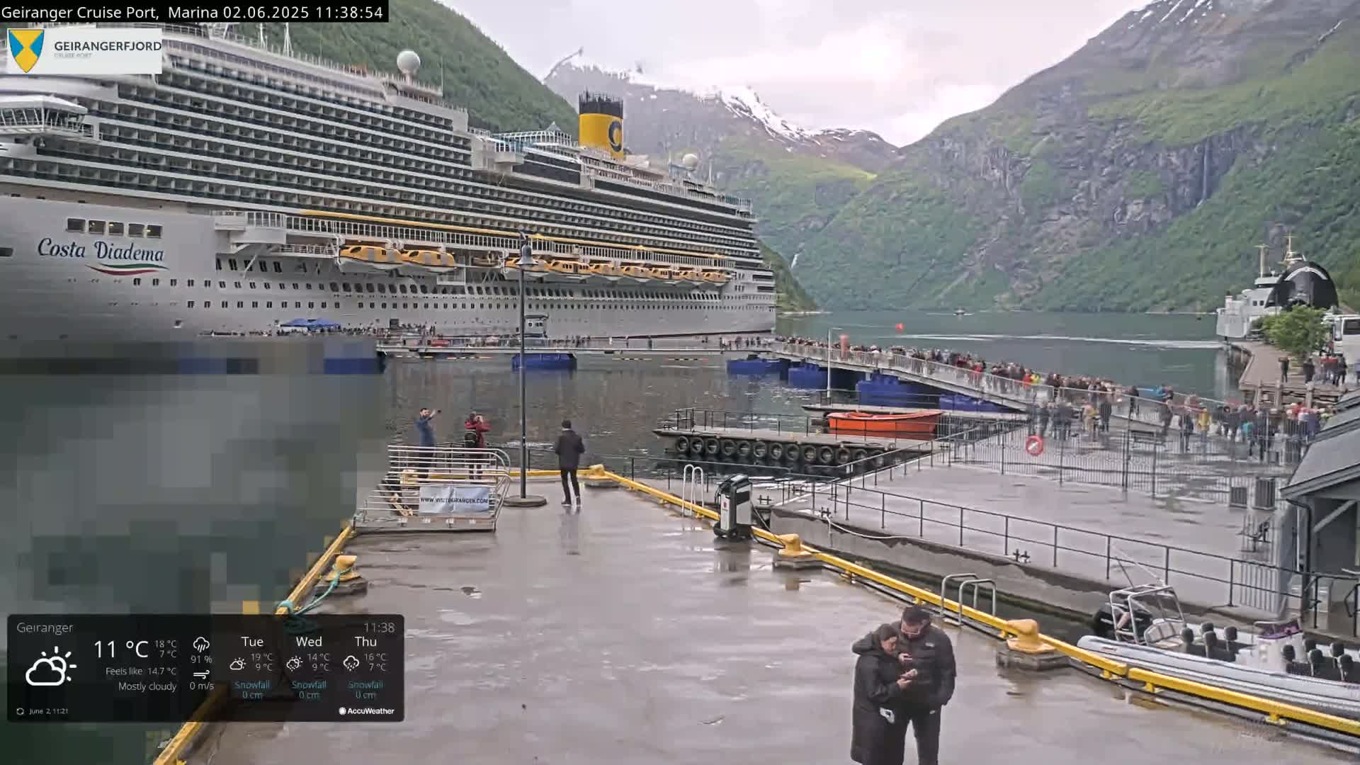 A large cruise ship is docked at a port in a fjord, surrounded by mountains and overcast weather, with many people gathered on the dock and nearby.