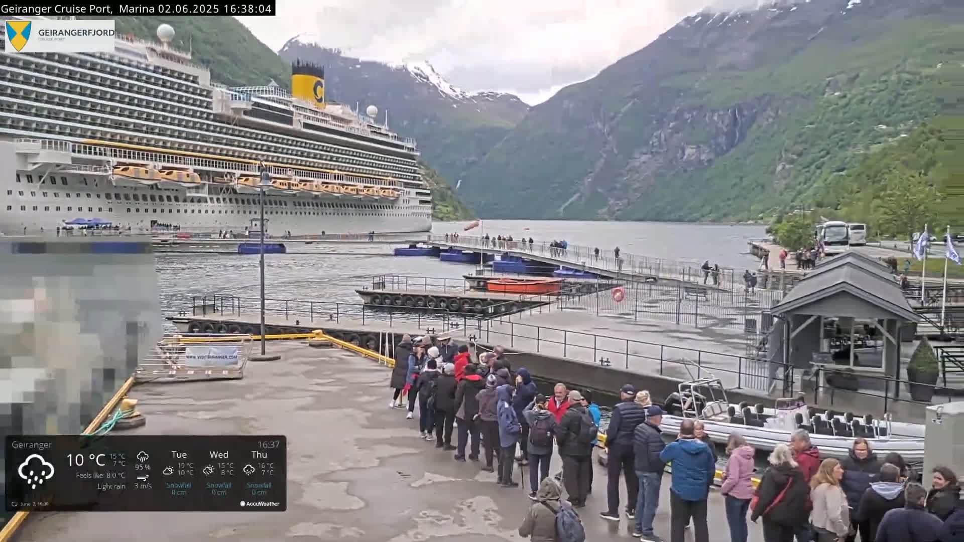 A large cruise ship is docked at a port in a fjord, surrounded by mountains under overcast, rainy conditions with a group of people waiting nearby.