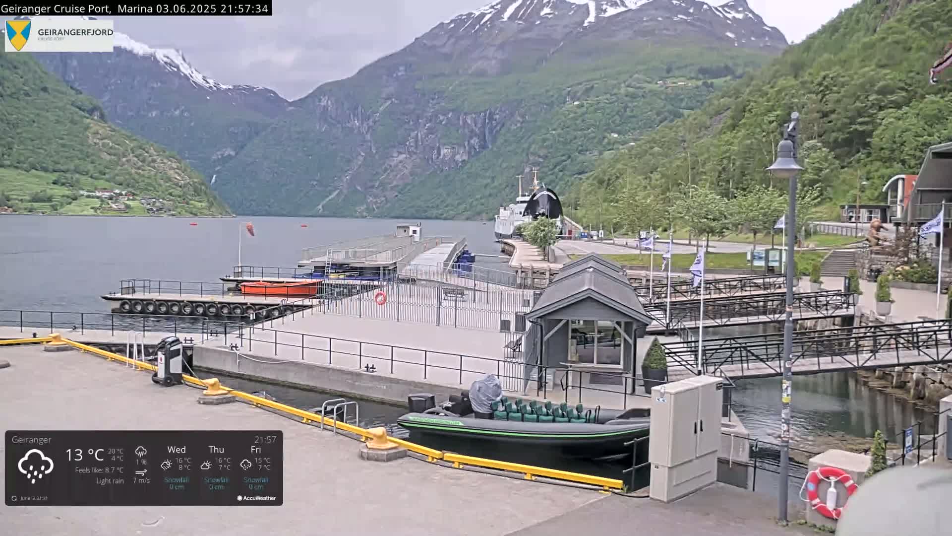 A marina with a docked boat and a small building is situated on a calm body of water, backed by snow-capped mountains under a lightly rainy sky.