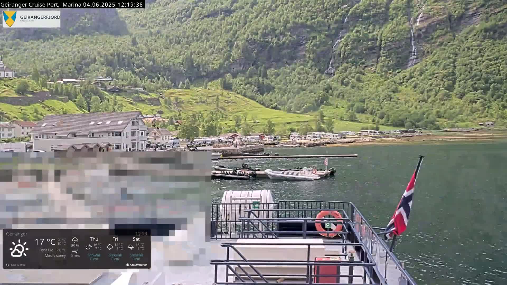 A mostly sunny view from a boat shows a calm fjord with several smaller vessels moored at a dock in front of a village nestled against a lush green mountainside.