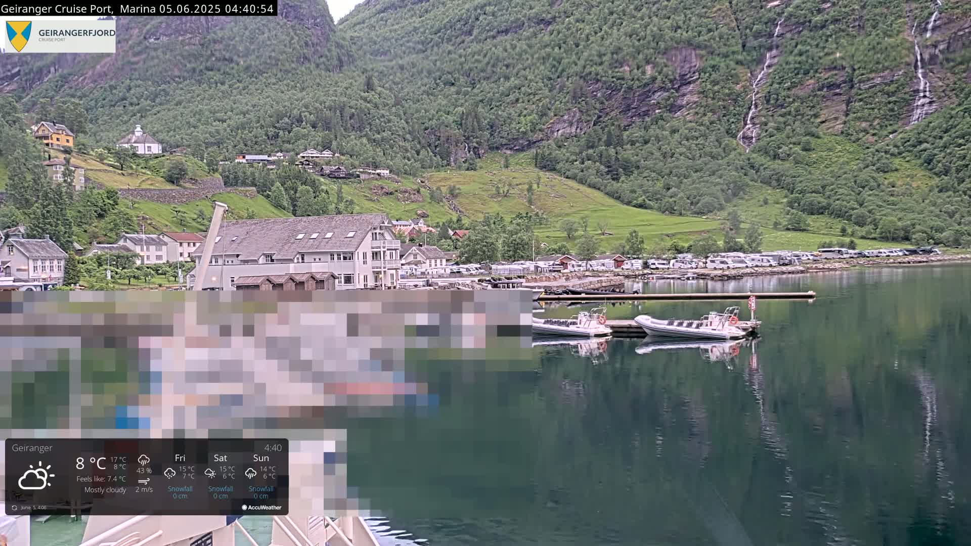 A mostly cloudy day reveals a calm fjord with a small town nestled at the base of steep, green mountains, several boats docked at a pier, and numerous RVs parked nearby.