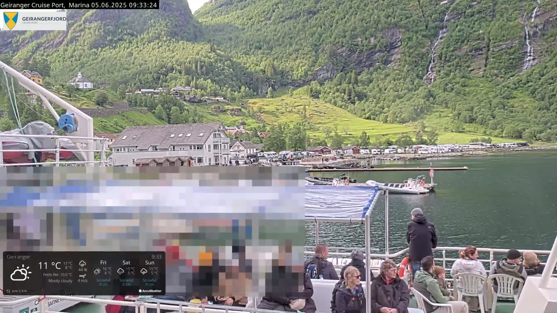 A mostly cloudy day in a fjord, with a ferry full of passengers moored near a small village nestled at the base of green mountains with several waterfalls cascading down their sides.