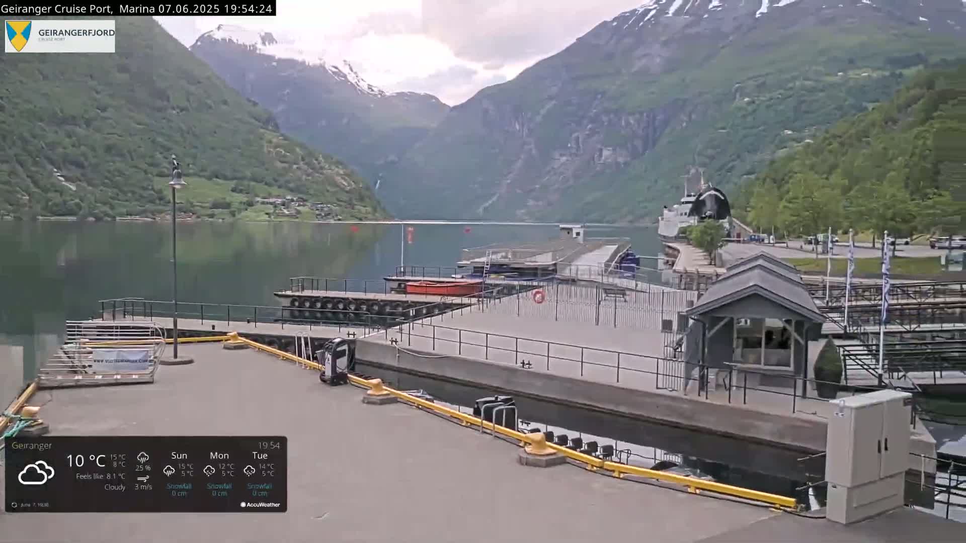 A cruise port situated on a calm fjord, surrounded by steep, green mountains under a cloudy sky.