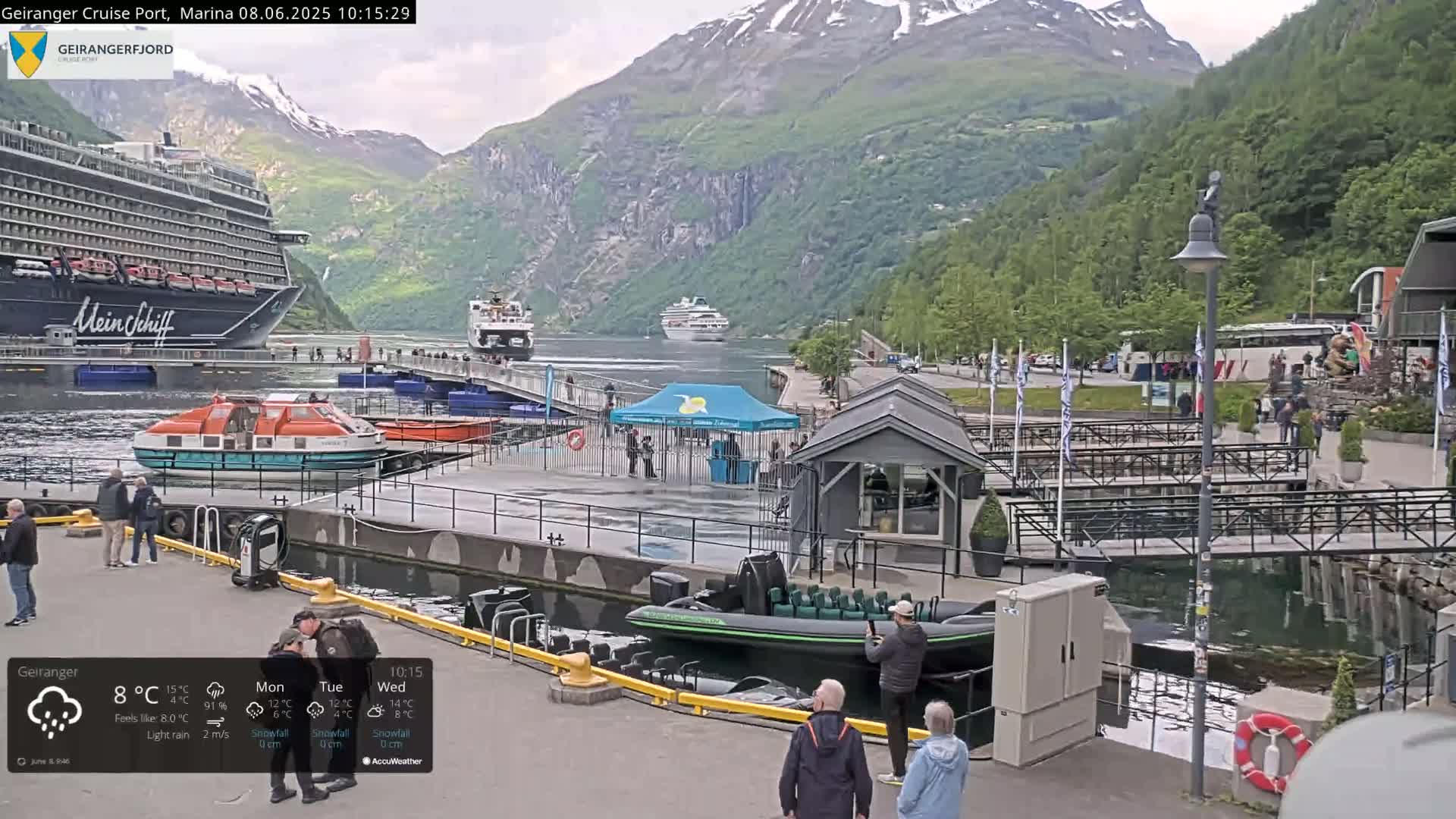 A cruise ship is docked at a marina in a fjord, surrounded by smaller boats, people, and buildings, under light rain and mountainous terrain.