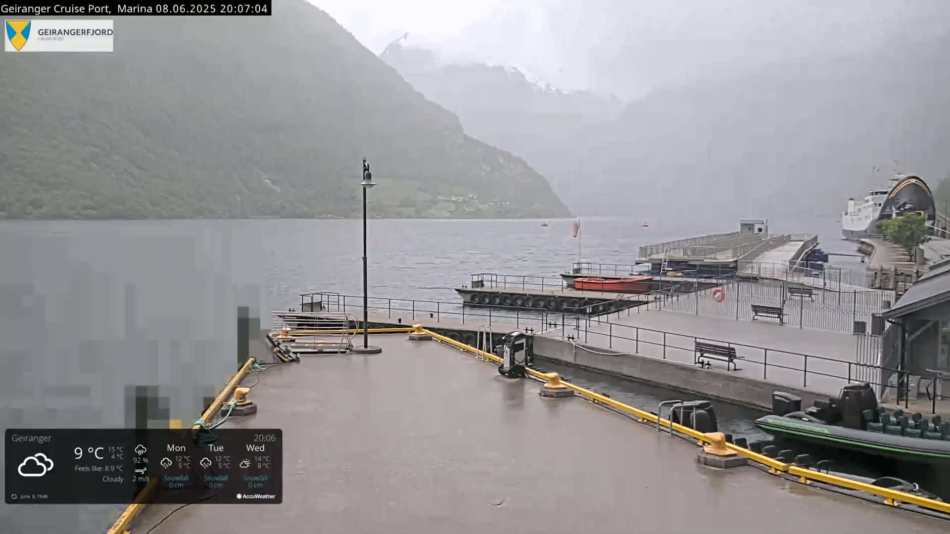 A rainy view of a harbor with boats docked, a ferry in the distance, and mountains in the background.