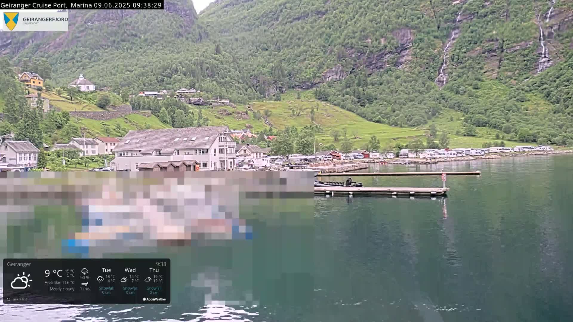 A mostly cloudy day shows a calm fjord with a small village nestled against steep, green mountains, featuring several boats docked at a marina.
