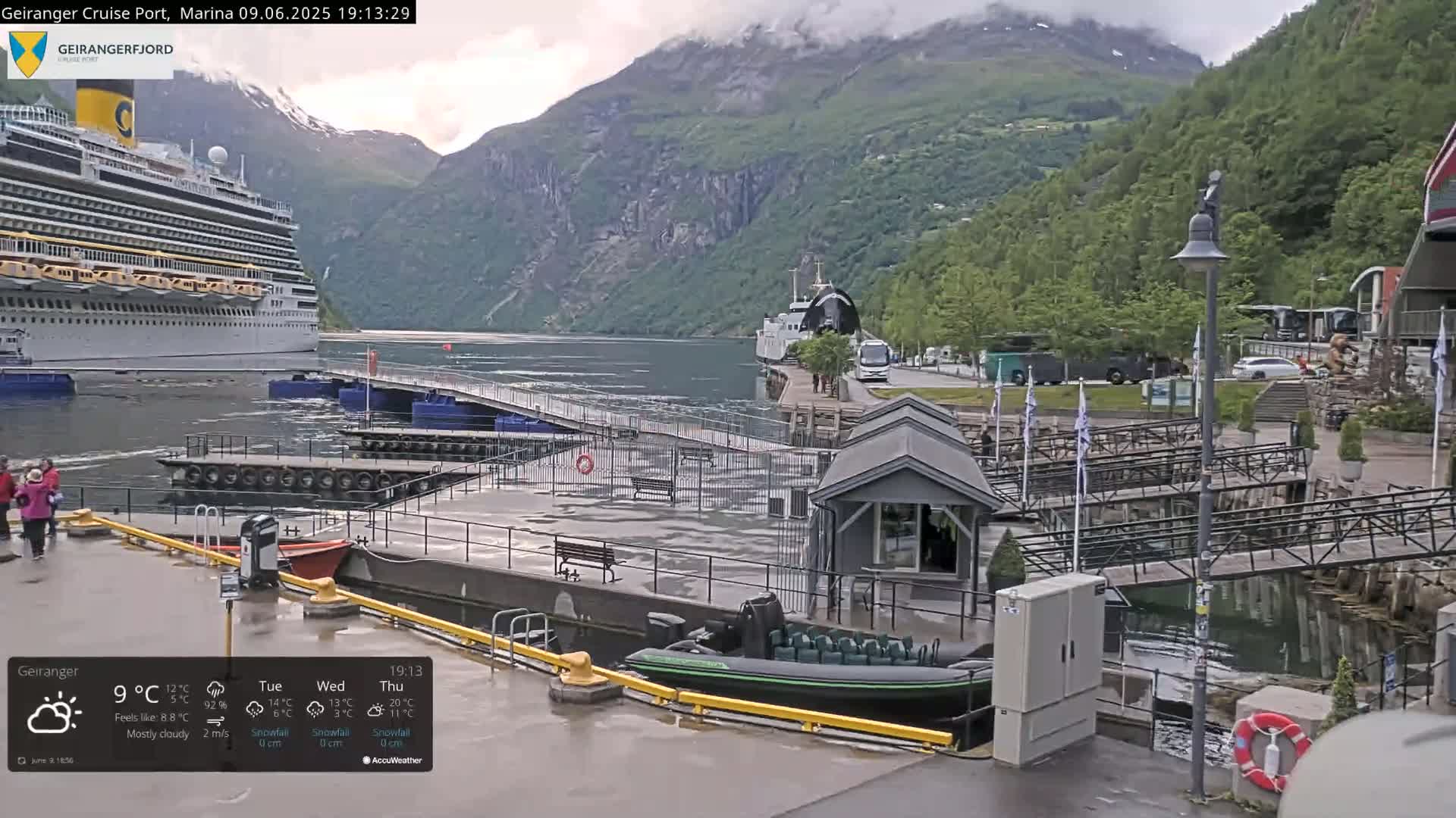 A cruise ship is docked at a port in a fjord surrounded by mountains under mostly cloudy skies.