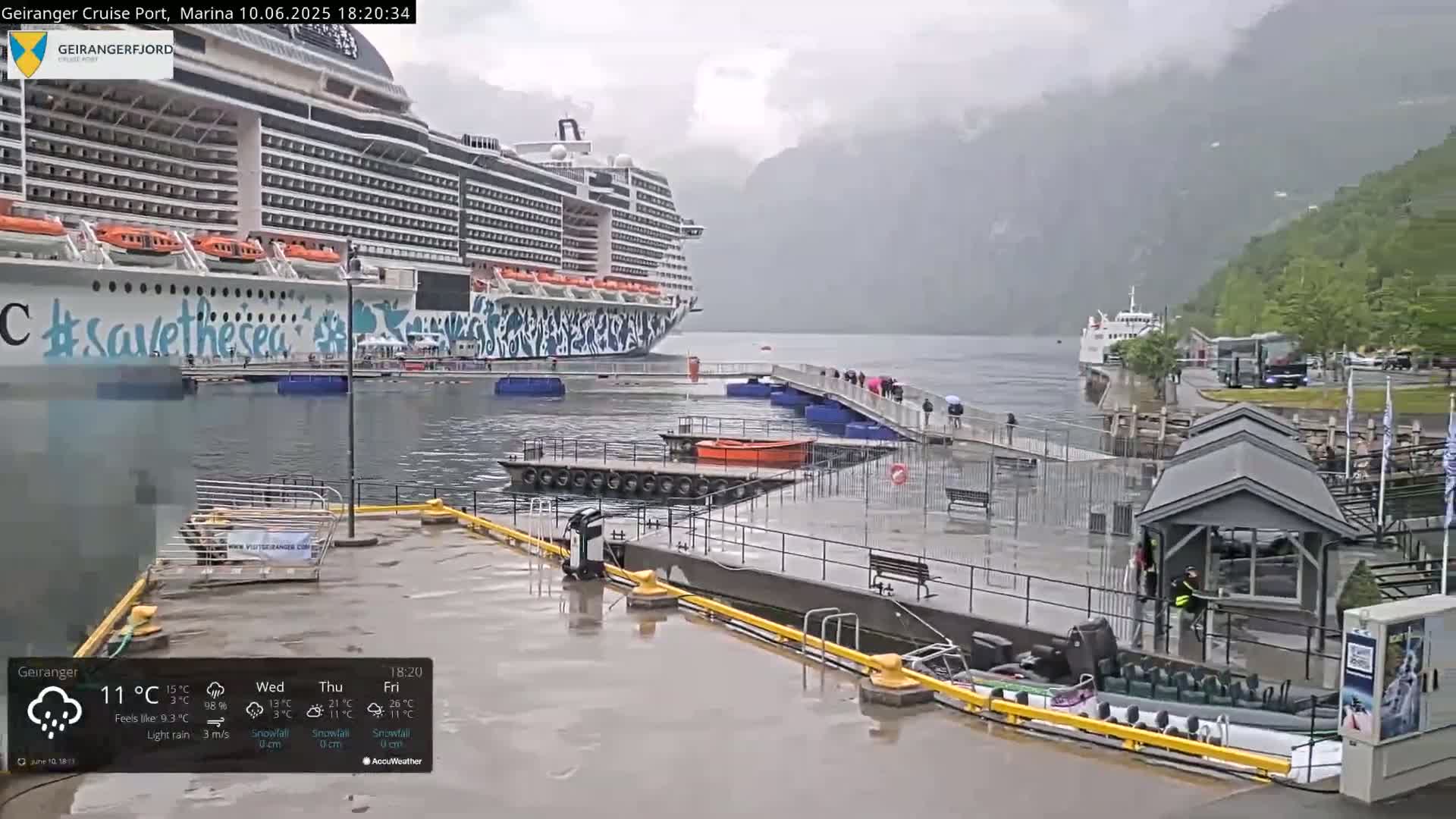 A large cruise ship is docked at a port in rainy weather, with several people walking along a pier and smaller boats moored nearby.