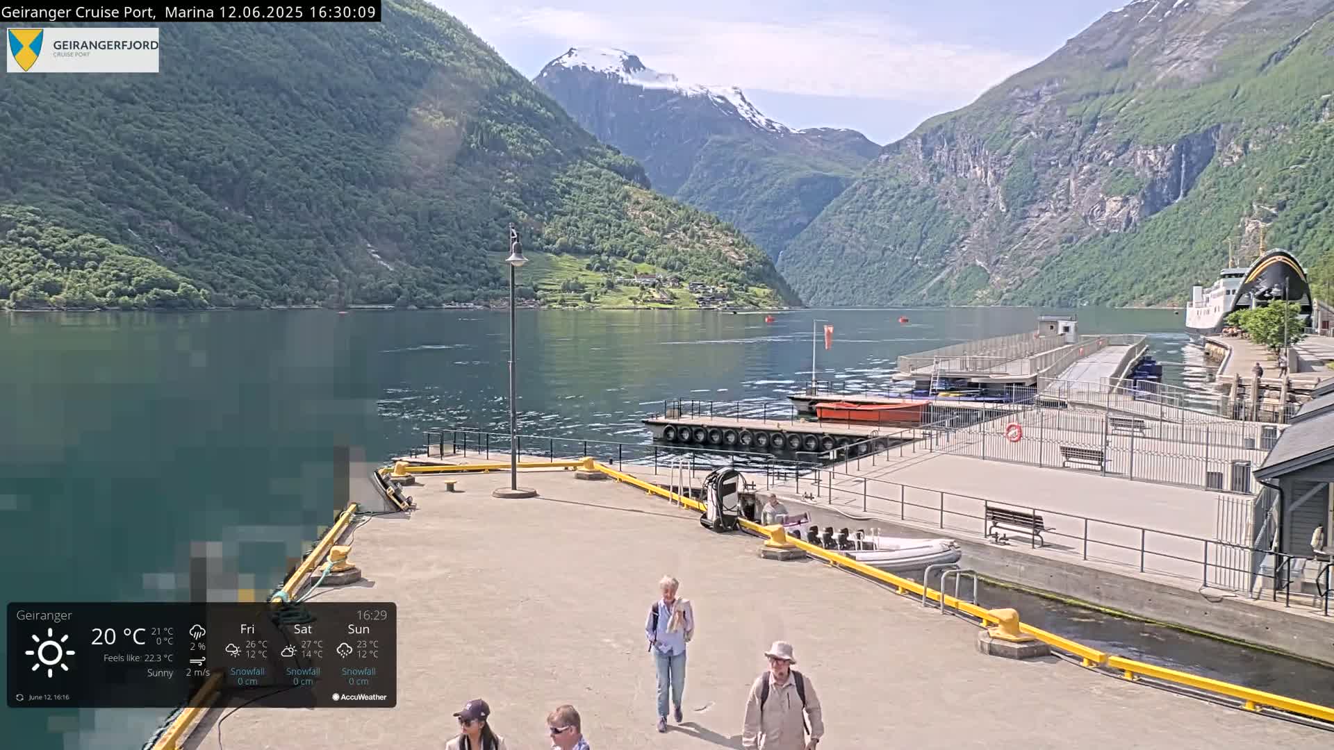 A sunny day at a cruise port shows a calm fjord surrounded by steep, green mountains, with a ferry docked and several people walking along the pier.