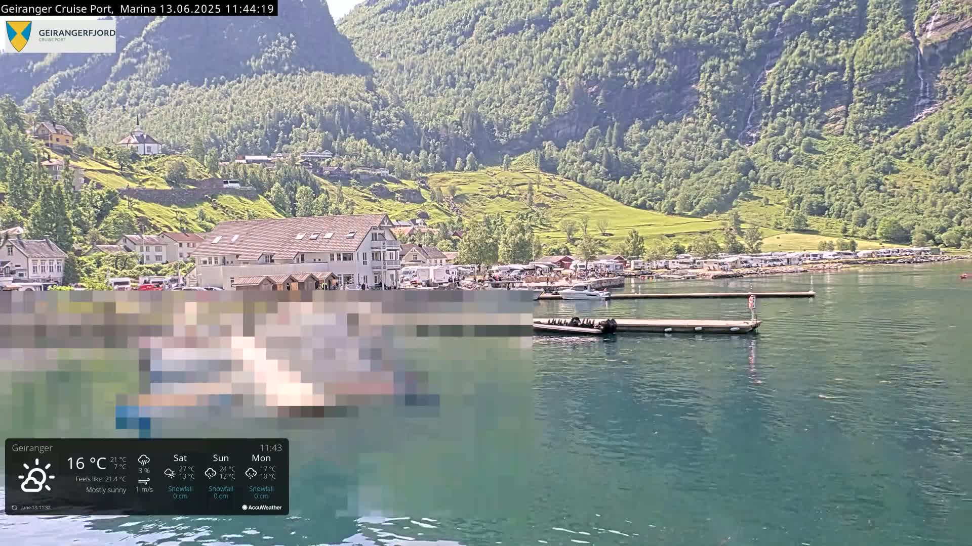 A mostly sunny waterfront village nestled against steep, green mountains, with boats moored at a dock.