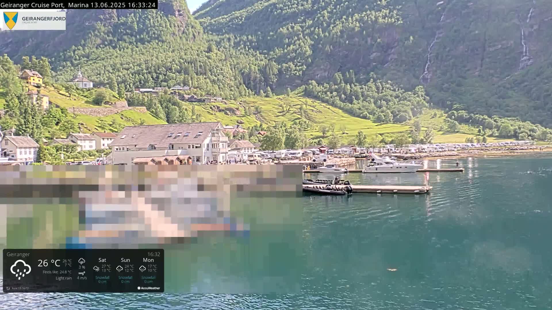 A calm fjord with several boats docked near a small town nestled against a steep, green mountainside under light rain.