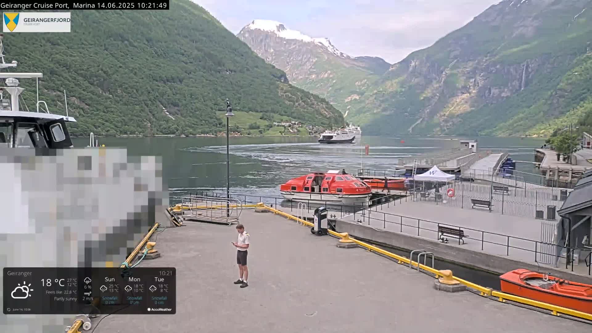 A partly sunny view of a cruise ship port nestled between steep, green mountains, with a lifeboat and smaller boats docked nearby.